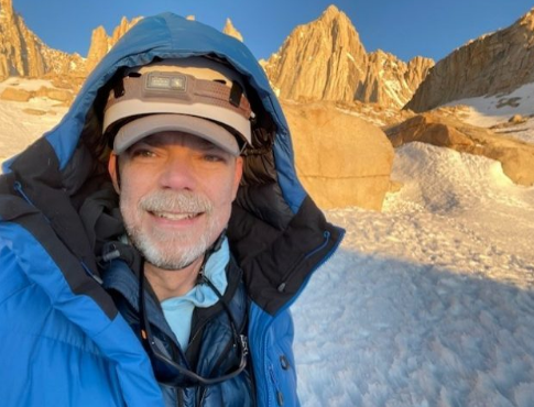 身穿蓝色外套、戴着帽子和头灯的微笑男子站在雪地里，身后是山峰。